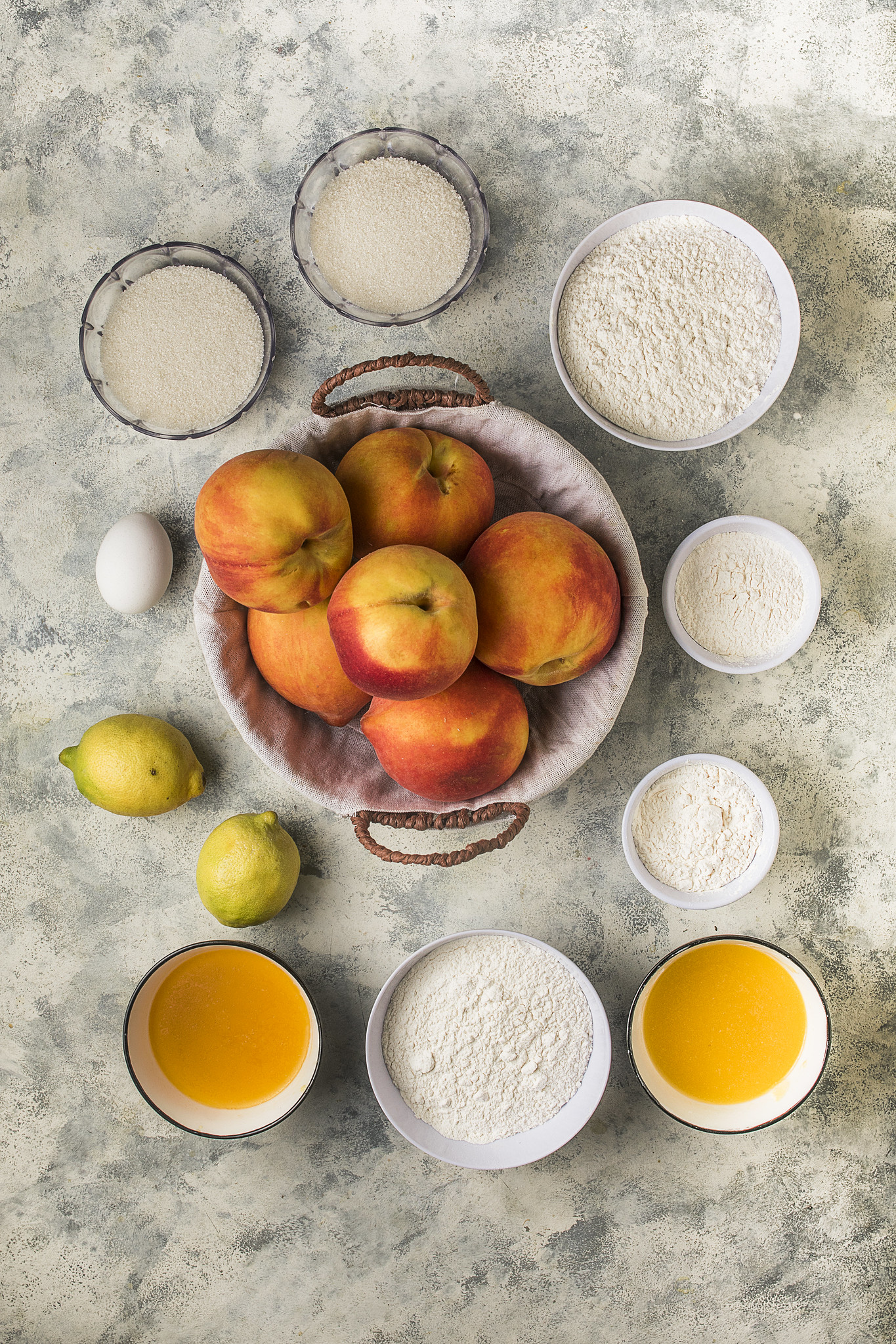 Overhead ingredient shot for sbriciolata di pesche, with fresh peaches, lemons, flour, cornflour, sugar, egg and melted butter arranged on a lightly textured surface.